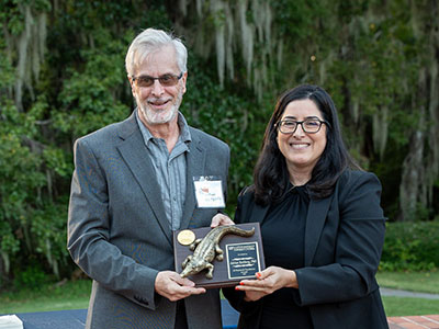 Two people holding an award plaque featuring a small alligator sculpture